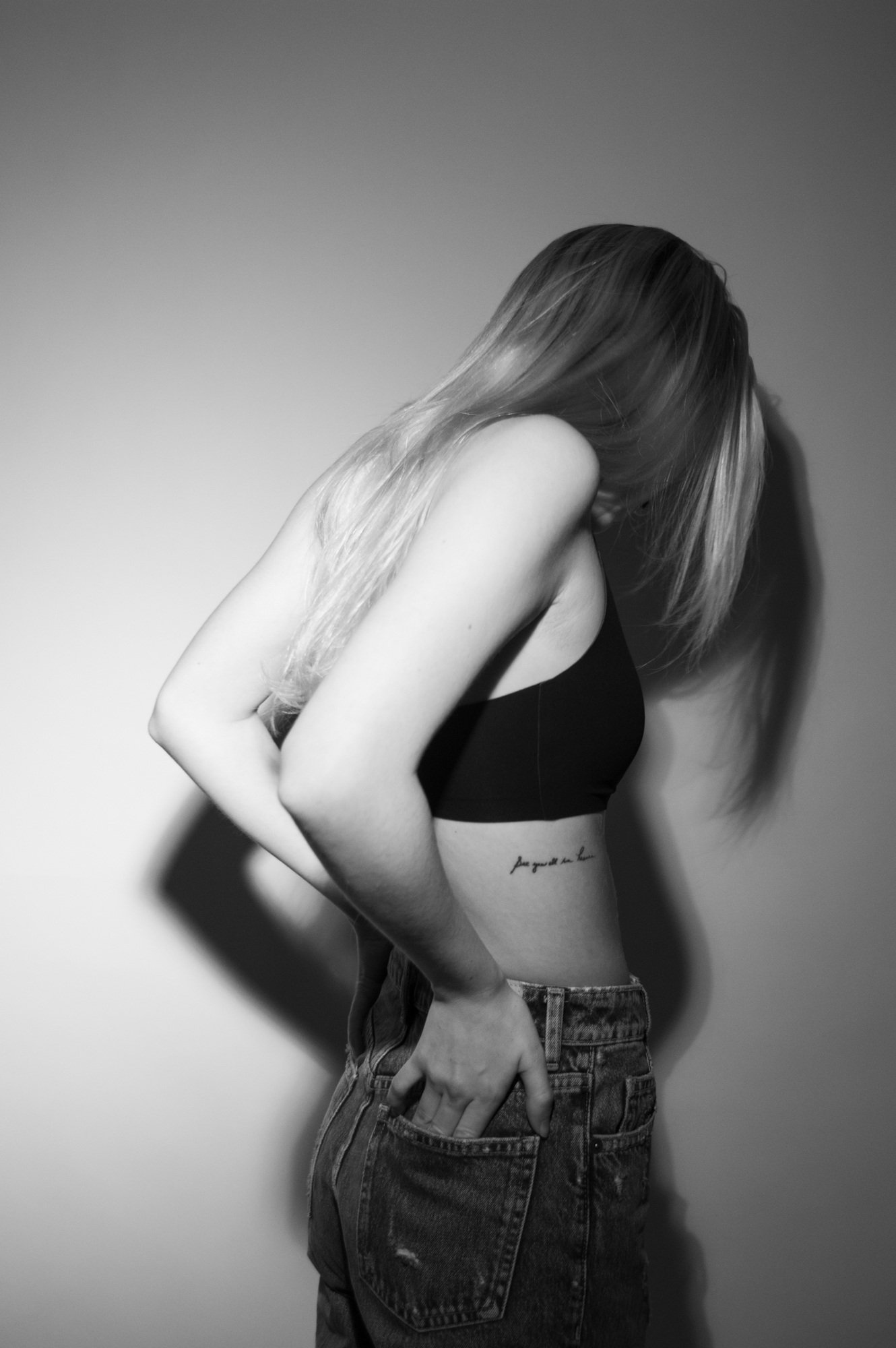Black and white image of the back of a slender blond female's head, so her hair is visible falling off of her shoulder. Small scripted tattoo visible on her side.