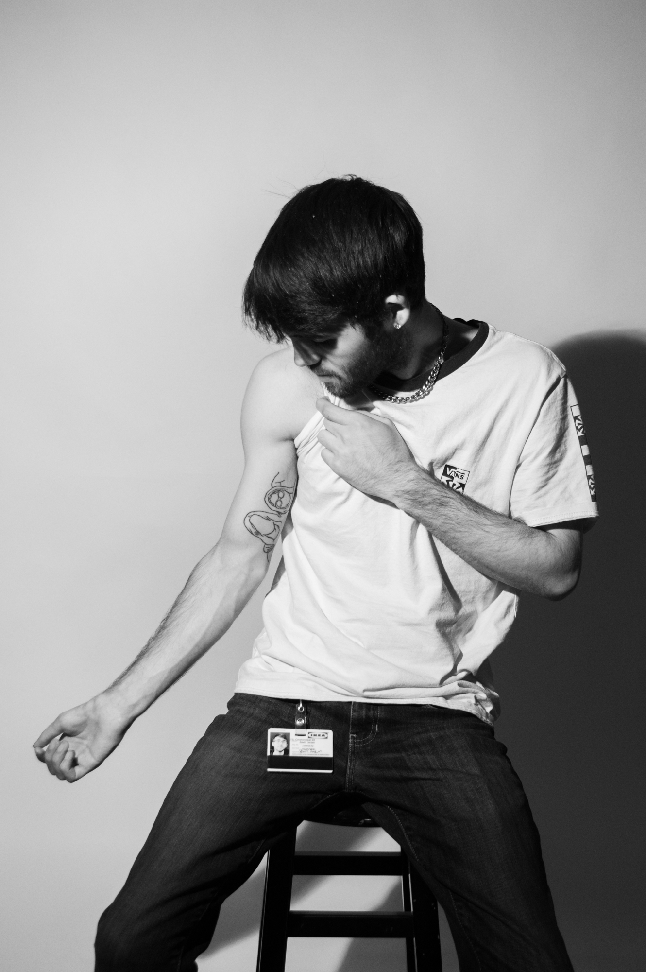 Black and white image of a white male with dark hair holding his arm out to the side to reveal a tattoo on the inside of his bicep. He is sitting on a stool.
