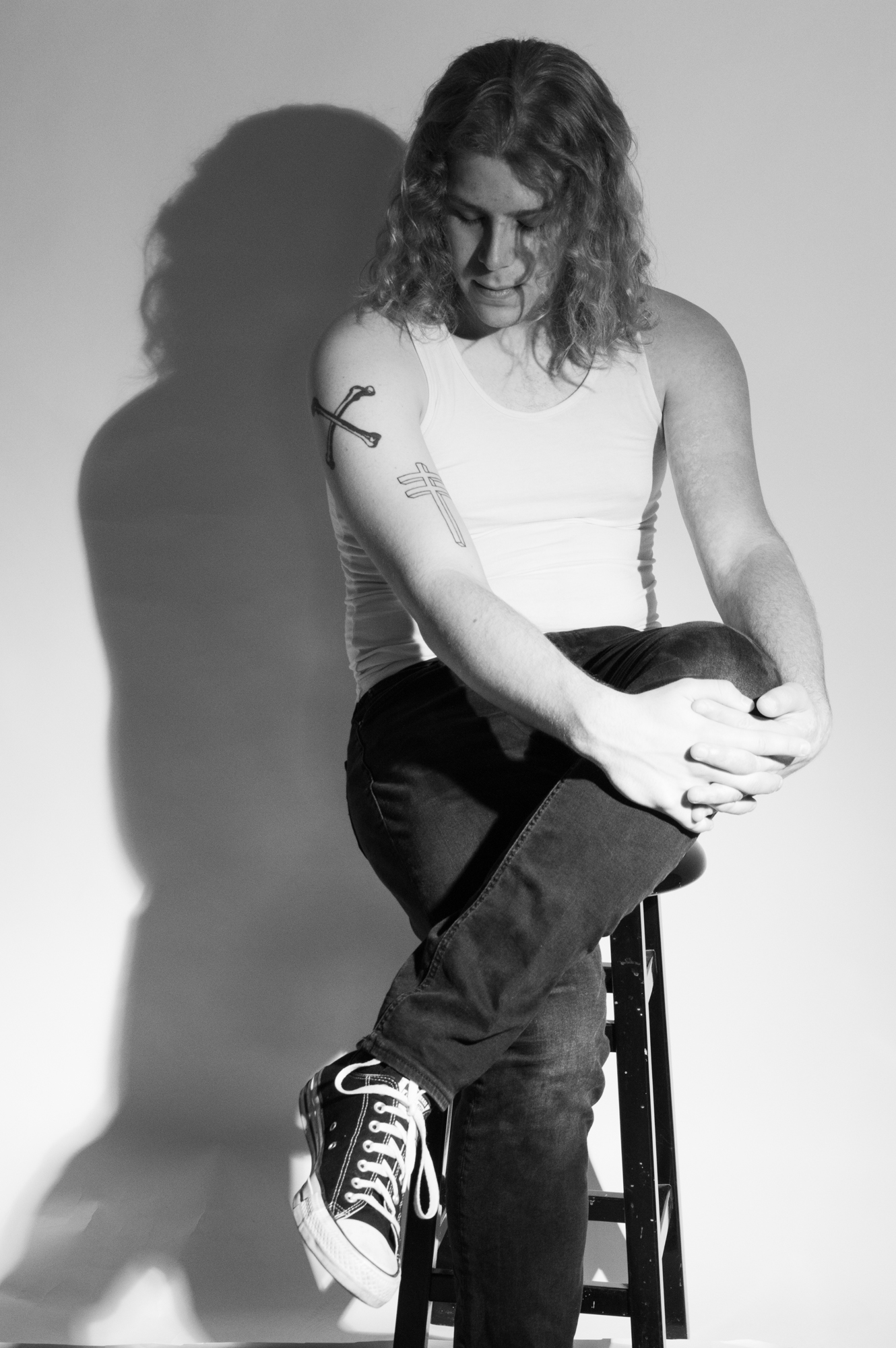 Black and white image of a tall, long-haired white male sitting on a stool with his legs crossed. Two tattoos visible on his right arm.