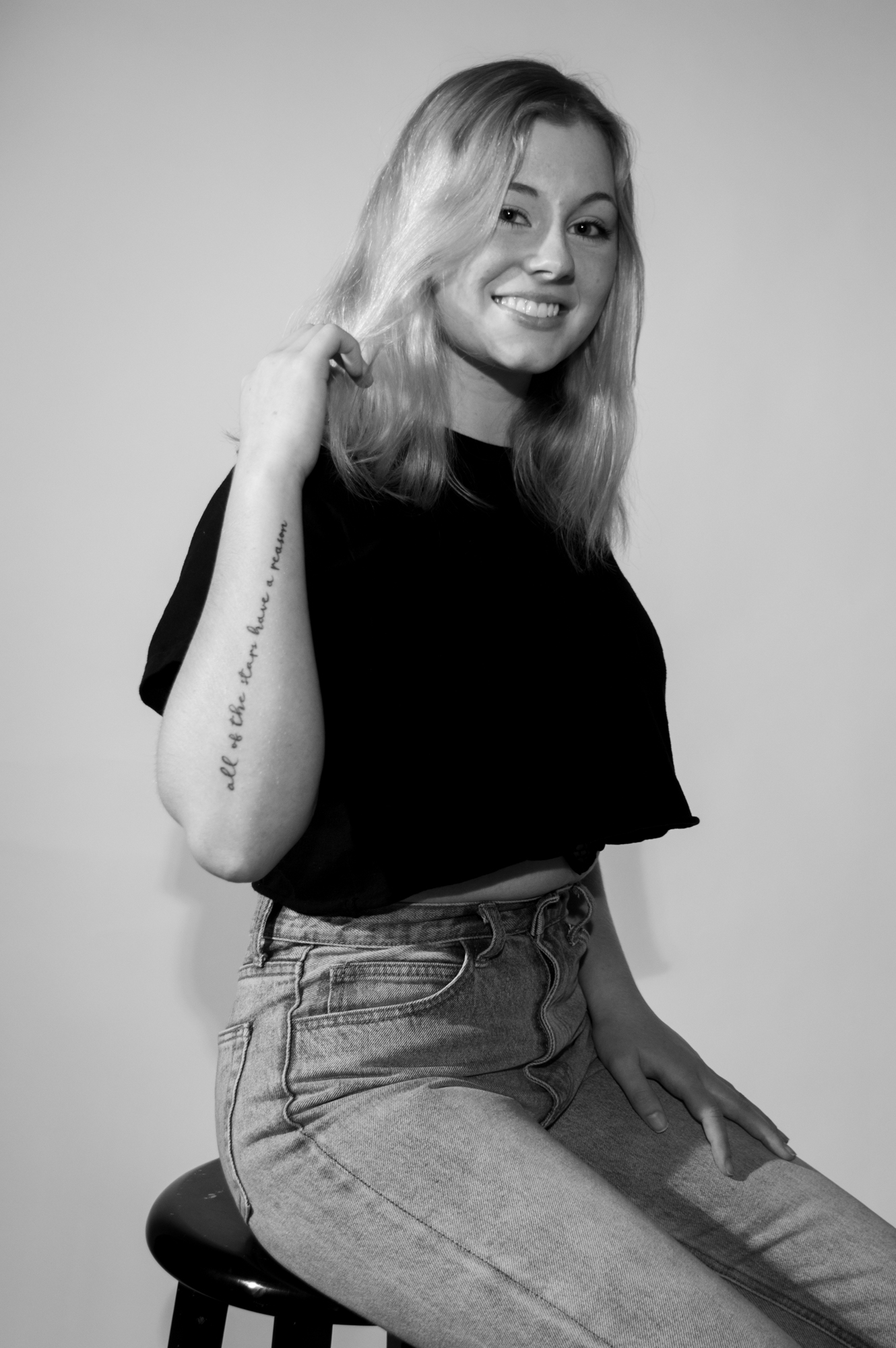 black and white image of a blond, white female sitting on a stool with a scripted tattoo on her forearm