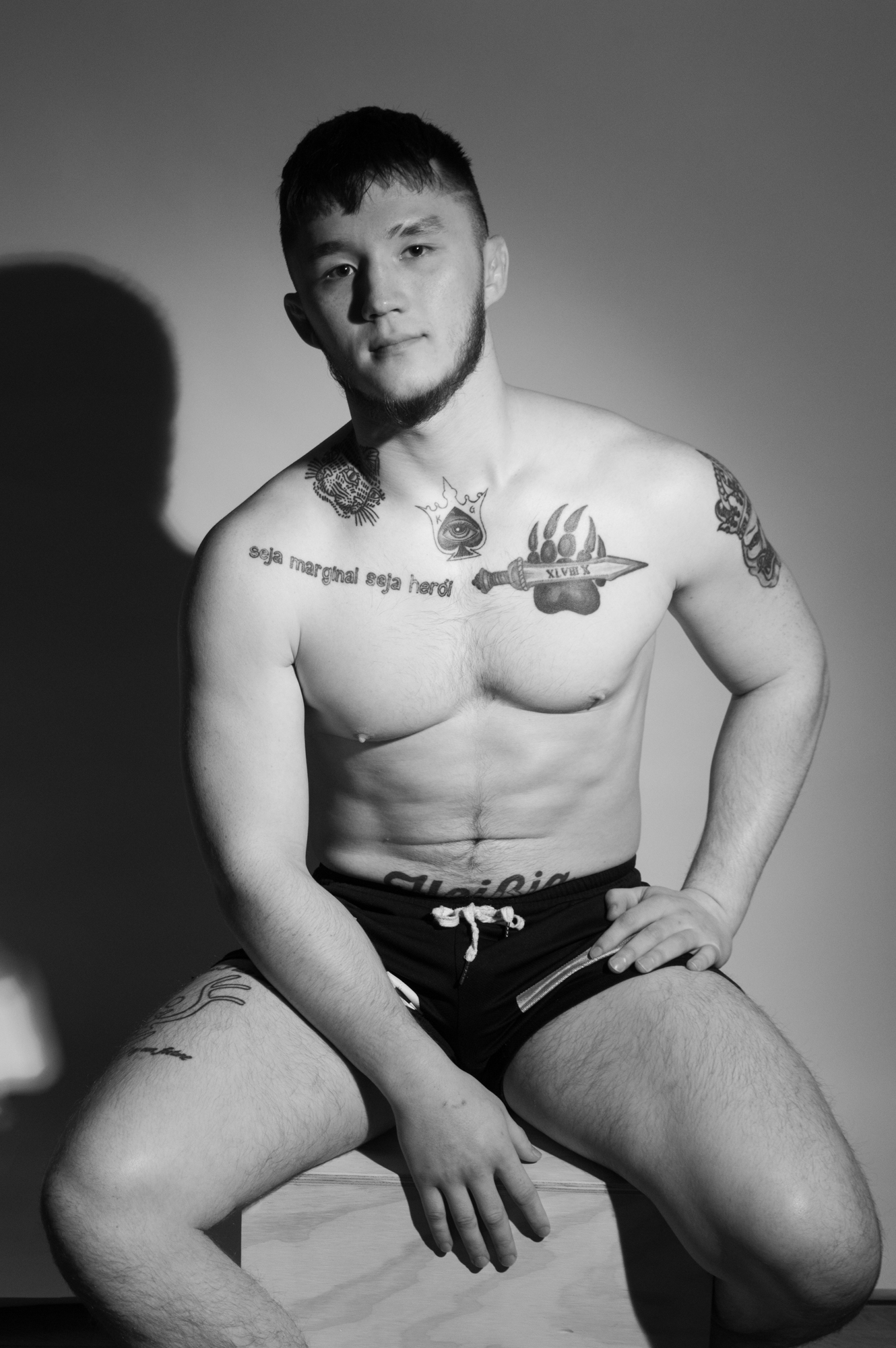 Black and white dramatic image of a male sitting on a wooden box with athletic build and beard with tattoos on his neck, chest, arm, leg and stomach.